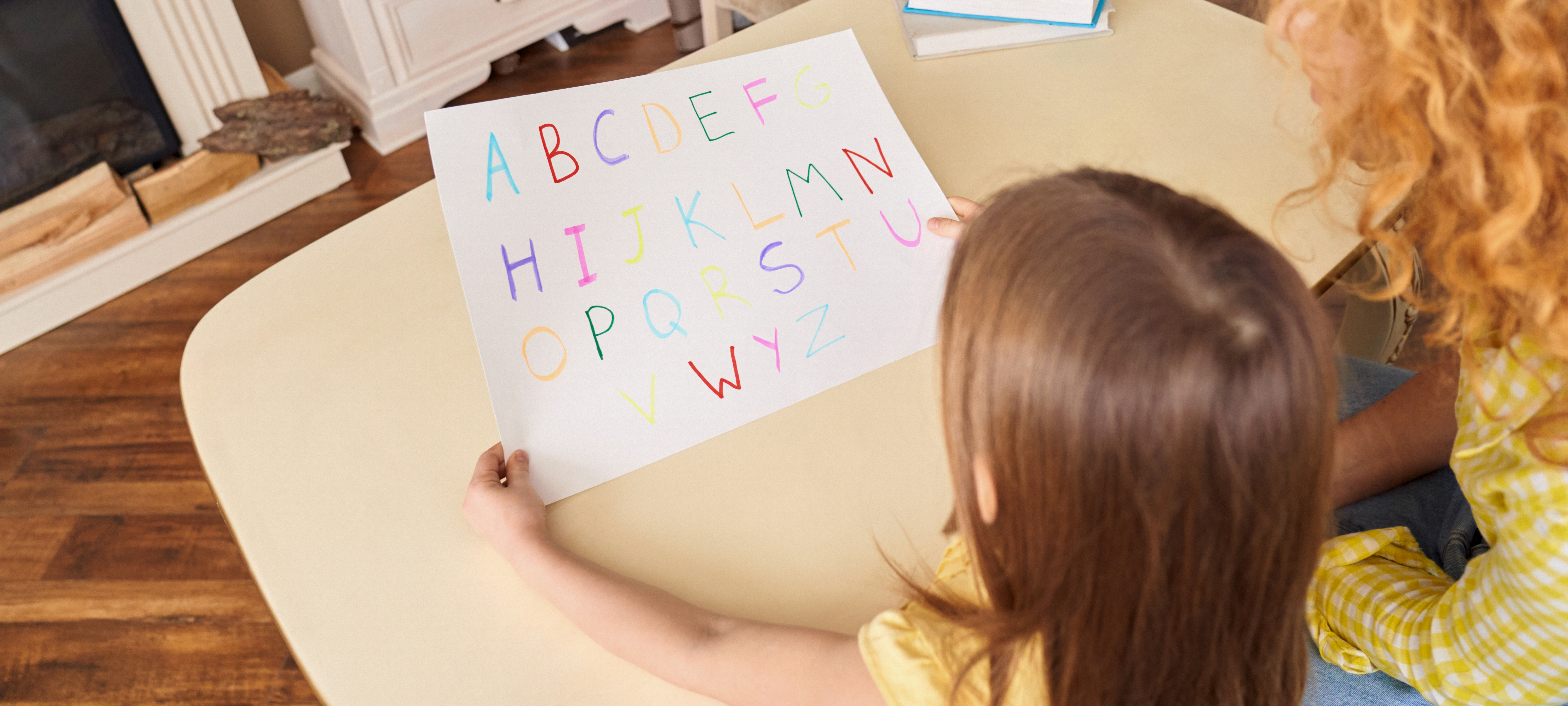 Tutor and student concept. Panoramic view of adult mother helping to her little daughter learning alphabet while they sitting behind desk, making home task for school together